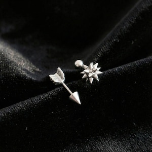 Könnte beinhalten: Zwei silberne Ohrringe auf schwarzem Hintergrund. Ein Ohrring ist als Pfeil gestaltet, der andere hat eine sternförmige Form. Der Pfeil-Ohrring hat eine Spitze und ein gefiedertes Ende. Der Stern-Ohrring hat mehrere spitze Kanten.