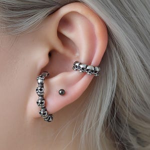 Könnte beinhalten: Silberner Ohrschmuck mit Totenkopf-Motiv. Das Bild zeigt einen Creolen-Ohrring mit mehreren Totenköpfen und einen Ohrklemmen-Ohrring mit einer Reihe von Totenköpfen. Ein kleiner, dunkler Ohrstecker ist ebenfalls sichtbar. Der Schmuck wird am Ohr einer Person getragen.