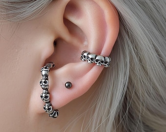 May include: Silver-coloured skull-themed ear jewellery. The image shows a hoop earring with multiple skulls and a cuff earring with a row of skulls. A small, dark stud earring is also visible. The jewellery is worn on a person's ear.