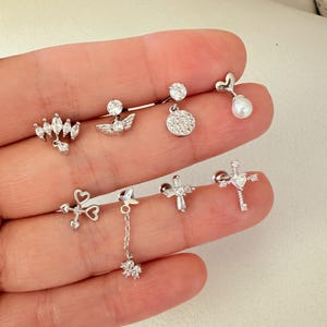 Puede incluir: Una colección de pendientes plateados con varios diseños. Los estilos incluyen un grupo en forma de marquesa, alas de ángel, un disco redondo, un corazón con una perla, un corazón y una flecha, una flor colgante, una cruz y una cruz con una gema en forma de corazón.