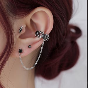 May include: Silver-toned ear cuff with a floral design and a dangling chain, paired with a black stud earring and a small black gemstone earring. The jewelry is worn on a person's ear.