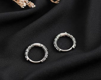 May include: A pair of silver hoop earrings with an engraved pattern, displayed on a dark cloth. The earrings are circular, featuring a detailed design around the circumference. The pattern consists of repeating shapes.