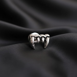Puede incluir: Un anillo plateado con forma de colmillos de vampiro. El anillo tiene una superficie detallada y texturizada, sobre un fondo oscuro y sedoso. Los colmillos son afilados y prominentes, creando un tema gótico o de Halloween.