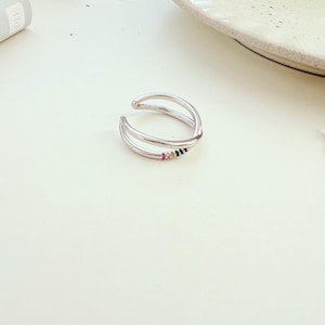 Puede incluir: Un anillo plateado de doble banda con una fila de pequeñas piedras preciosas de colores. El anillo tiene un diseño abierto, lo que permite ajustarlo. Las piedras preciosas están en un patrón de arcoíris, agregando color al diseño.