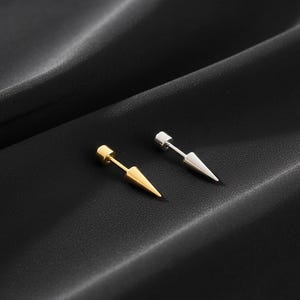 Puede incluir: Dos pendientes de botón con un diseño de punta. Un pendiente es de color dorado y el otro es de color plateado. Los pendientes tienen un poste y una punta cónica. Los pendientes se muestran sobre una superficie negra texturizada.