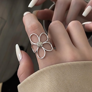 Może przedstawiać: Srebrny pierścionek z motywem kwiatowym. Pierścionek ma pięć ażurowych płatków otaczających teksturowane, złote centrum. Pierścionek jest noszony na palcu, prezentując swój delikatny kwiatowy kształt.