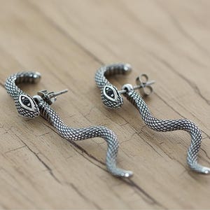 May include: A pair of silver-toned snake earrings. Each earring features a snake head with black eye detailing and a textured body that curves into a stylized S-shape. The earrings are set against a wooden background.