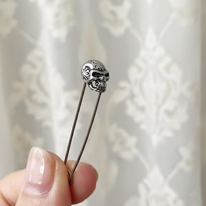 Puede incluir: Un alfiler plateado con forma de calavera y detalles intrincados. La calavera está montada sobre un alfiler recto, creando un accesorio único. El alfiler se muestra sobre un fondo suave y borroso, resaltando el diseño de la calavera.