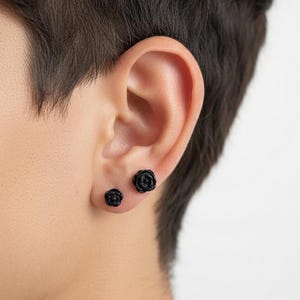 May include: Black rose-shaped stud earrings. The earrings are small and detailed, with a textured surface mimicking the petals of a rose. The earrings are shown on an ear, with one earring slightly smaller than the other.