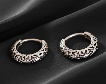 May include: Two silver hoop earrings with an ornate, repeating pattern. The earrings are displayed on a dark, textured surface, which accentuates their detailed design and metallic finish. The earrings are a classic jewellery item.