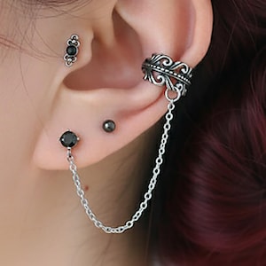 Puede incluir: Conjunto de joyas para la oreja con un pendiente de botón de piedra negra, un pendiente de cadena y un ear cuff. El ear cuff tiene un diseño ornamentado. El pendiente de cadena conecta el botón con el ear cuff.