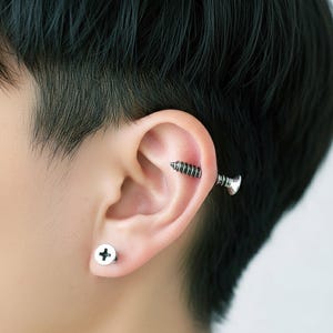 May include: An ear with two silver-coloured screw-shaped earrings. One is a stud, and the other is a barbell style that appears to go through the ear. The earrings are a unique and edgy accessory, a statement piece.