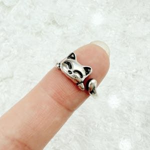 May include: A silver-colored ring shaped like a cat. The cat's head and paws wrap around the finger, with black accents on the ears and face. The ring is a cute and whimsical accessory.