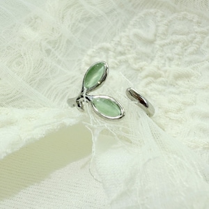 Puede incluir: Anillo plateado con dos gemas ovaladas de color verde pálido en un diseño de hojas. La banda ajustable es abierta. El anillo se encuentra sobre un fondo blanco texturizado. Las gemas son de color verde claro.