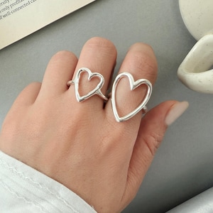 Könnte beinhalten: Zwei silberne herzförmige Ringe werden auf einer Hand gezeigt. Die Ringe haben ein minimalistisches Design, wobei die Herzform durch ein durchgehendes Metallband gebildet wird. Die Ringe werden an verschiedenen Fingern getragen und zeigen ihre Größe und ihren Stil.