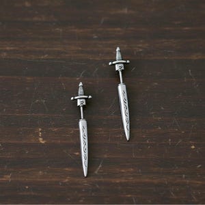 Puede incluir: Un par de pendientes de daga plateados. Cada pendiente presenta una empuñadura detallada y una hoja con diseños grabados. Los pendientes se exhiben sobre una superficie de madera marrón oscuro.