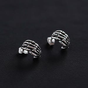 Könnte beinhalten: Ein Paar silberfarbene Ohrstecker. Jeder Ohrring ist wie eine Skeletthand geformt, mit detaillierten Fingerknochen. Die Ohrringe sind vor einem dunklen Hintergrund platziert, der das filigrane Design und den metallischen Glanz hervorhebt.
