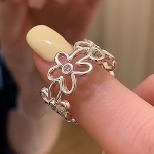 Puede incluir: Un anillo plateado con un diseño floral. Cada flor tiene cinco pétalos y una pequeña piedra transparente en el centro. El anillo se lleva en un dedo con esmalte de uñas amarillo.