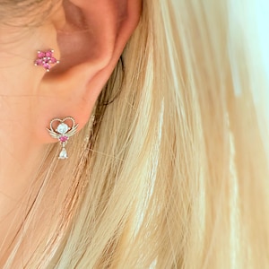 Może przedstawiać: Zbliżenie ucha ozdobionego dwoma kolczykami. Jeden to mały, różowy, w kształcie gwiazdy. Drugi to srebrny kolczyk z sercowatym wierzchem, skrzydłami i wiszącym klejnotem w kształcie łzy. Kolczyki są osadzone na tle blond włosów.