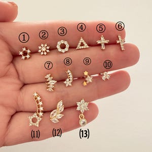 Può includere: Una collezione di tredici orecchini a bottone color oro con vari design, tra cui stelle, croci, fiori e forme geometriche. Gli orecchini sono adornati con piccole pietre preziose trasparenti, creando un effetto scintillante. Gli orecchini sono esposti su uno sfondo chiaro.