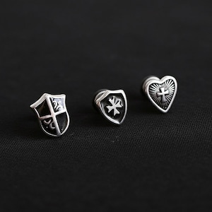 May include: Three silver-toned stud earrings with different designs. One is a shield shape with a black center and a cross design. Another is a shield shape with a star design. The third is a heart shape with a cross design.