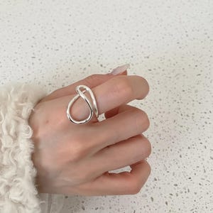 Può includere: Un anello color argento con un design astratto unico. L'anello presenta una forma continua e ad anello che avvolge il dito. L'anello è mostrato su una mano, su uno sfondo chiaro.