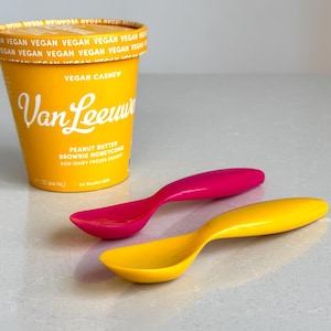 May include: A yellow container of vegan cashew ice cream with the brand name "Van Leeuwen" in white script. The container is next to a pink and a yellow spoon. The ice cream flavor is peanut butter brownie honeycomb.