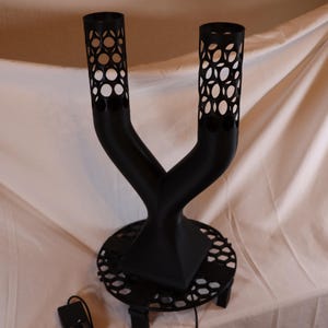 May include: A black, Y-shaped decorative object with two cylindrical tops featuring a pattern of circular cutouts. The object sits on a round base with a similar cutout design. A power cord and adapter are visible.
