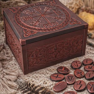 Peut inclure: Une boîte en bois marron avec des motifs sculptés complexes, dont une rose des vents sur le couvercle et des motifs de nœuds celtiques sur les côtés. Plusieurs petits jetons ronds, gravés de runes, sont dispersés à proximité.