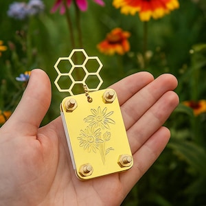 May include: A gold-colored, rectangular pendant with a honeycomb design and daisy engraving. The pendant has four rounded corner screws and is held in a hand against a blurred background of colorful flowers.