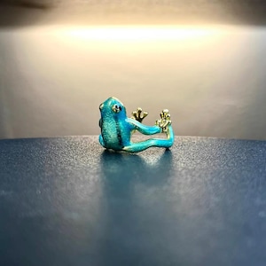 May include: A turquoise blue frog ring with gold accents. The frog is sitting on its hind legs with its front legs raised.