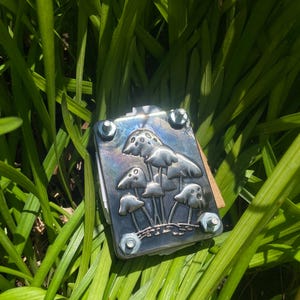 May include: A silver-colored metal notebook with a mushroom design. The notebook is secured with four screws and is surrounded by green grass.