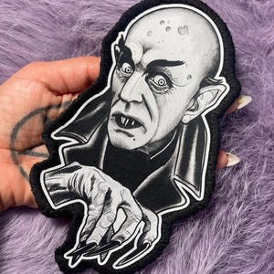 Puede incluir: Un parche bordado en blanco y negro con una ilustración detallada de un vampiro calvo de piel pálida, con orejas puntiagudas, dientes afilados y uñas largas como garras. La figura viste un abrigo oscuro con cuello alto y un borde blanco.