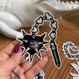 May include: Black and white sticker featuring a heart-shaped mace with spikes, connected by a chain of hearts to a cylindrical handle. The sticker has a tattoo-inspired design, with a detailed, shaded appearance.