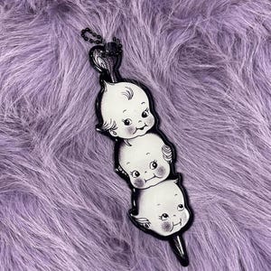Pode incluir: Um pingente preto e branco com três rostos de bebês sorrindo empilhados verticalmente. O pingente tem uma corrente preta e está sobre um fundo roxo e fofo. Os bebês têm bochechas rosadas e traços faciais detalhados.