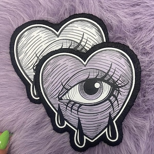 Puede incluir: Dos parches bordados en forma de corazón que presentan un ojo central con pestañas largas y tres lágrimas negras. Los parches tienen un borde negro y una base de color violeta claro o blanco con un detallado trabajo de líneas negras.