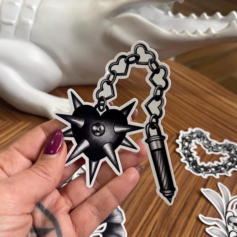 May include: Black and white sticker featuring a heart-shaped mace with spikes, connected by a chain of hearts to a cylindrical handle. The sticker is held in a hand with a dark purple nail polish.