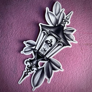 Peut inclure: Autocollant noir et blanc représentant une lanterne vintage détaillée avec un serpent et une bougie à l'intérieur. La lanterne est entourée de feuilles et possède un fleuron décoratif. L'autocollant est posé sur un fond violet texturé.