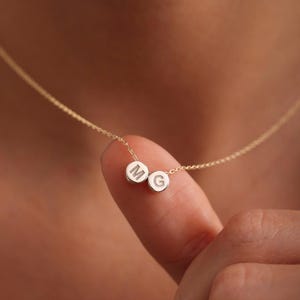 Può includere: Una delicata collana d'oro con due piccoli ciondoli rotondi. Ogni ciondolo ha un'iniziale incisa, con le lettere "M" e "G" visibili. La collana è mostrata su uno sfondo sfocato.