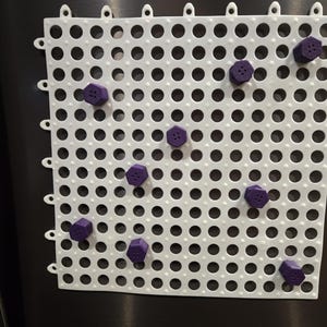 May include: A white, perforated plastic mat with evenly spaced holes and a series of small loops along the top edge. Scattered across the mat are several purple, hexagonal-shaped magnets, each with a small, circular indentation.