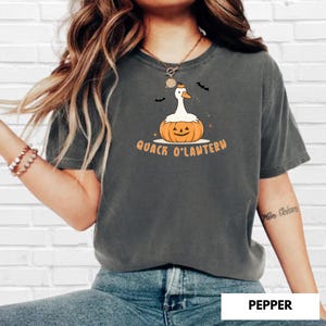 May include: Dark grey t-shirt with a Halloween-themed graphic. The design features a white duck with an orange beak and a pumpkin body, with the text "Quack O'Lantern" in orange. Two black bats are above the duck.
