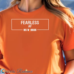 May include: Orange t-shirt with the text "FEARLESS AF MS WARRIOR" in a white rectangular box. The shirt has short sleeves and a crew neck. The text is in a bold, sans-serif font. The shirt is a solid orange color.