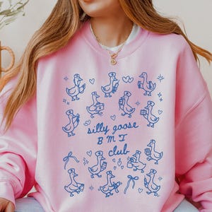 May include: A light pink sweatshirt featuring a blue illustration of geese wearing nurse hats and carrying medical supplies. The text "silly goose BMI club" is printed below the geese. The sweatshirt is a crew neck style.