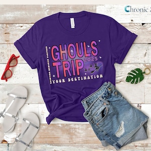 May include: A purple t-shirt with the words "Ghouls Trip 2025" in a gradient font, along with a witch graphic. The shirt is paired with denim shorts, red glasses, and silver sandals. A green leaf keychain is also present.