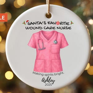 May include: A round, white Christmas ornament with a gold string. The ornament features a pink medical scrub top with a stethoscope, and the text "Santa's Favorite Wound Care Nurse". Below the scrub top is the text "Making spirits bright. Ashley 2025".