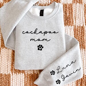May include: A light gray sweatshirt with the words "cockapoo mom" and a paw print graphic on the front. The sleeve features the names "Luna" and "Gavin" with paw prints. The sweatshirt is made of a soft material.