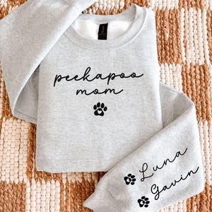 May include: A light gray sweatshirt with the words "peekapoo mom" and a paw print graphic on the front. The sleeve features the names "Luna" and "Gavin" with paw prints. The sweatshirt is made of a soft material.