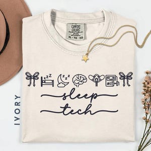 May include: Ivory-colored t-shirt with the words "sleep tech" in a script font. The shirt features black line art of a bow, bed, moon, brain, oxygen mask, and a machine. A gold star pendant necklace is draped on the shirt.