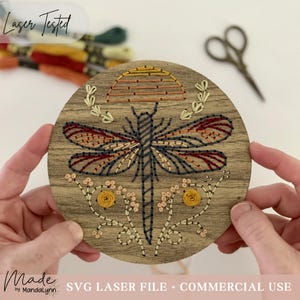 Puede incluir: Una placa de madera circular con un diseño de libélula bordado. La libélula tiene alas cosidas en azul oscuro y rojo. Arriba hay un motivo de sol semicircular en amarillo y naranja. Debajo hay tallos florales con nudos amarillos.
