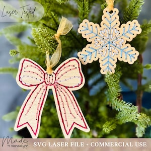 May include: Two wooden Christmas decorations hang from a green tree. One is a bow shape with red and pink stitching, the other is a snowflake with blue and white stitching. The ornaments are laser cut and have a light wood colour.